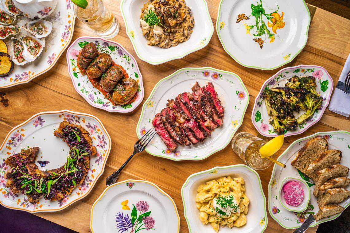 A high-angle, overhead flat lay of a large, family-style feast spread across a wooden table. A platter of sliced, medium-rare steak sits in the center, surrounded by numerous side dishes including macaroni and cheese, charred broccoli, oysters, roasted potatoes, and a large pork chop. The food is served on decorative, floral-patterned plates, with several drinks also on the table.