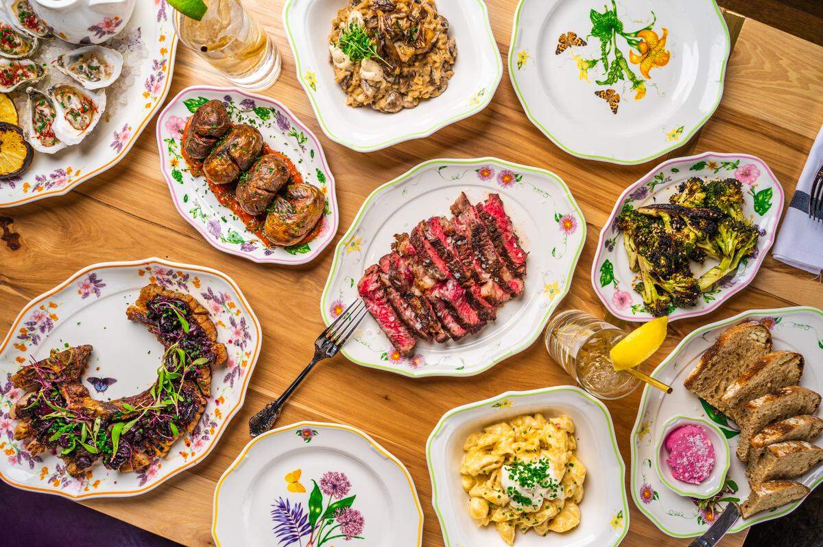 A high-angle, overhead flat lay of a large, family-style feast spread across a wooden table. A platter of sliced, medium-rare steak sits in the center, surrounded by numerous side dishes including macaroni and cheese, charred broccoli, oysters, roasted potatoes, and a large pork chop. The food is served on decorative, floral-patterned plates, with several drinks also on the table.
