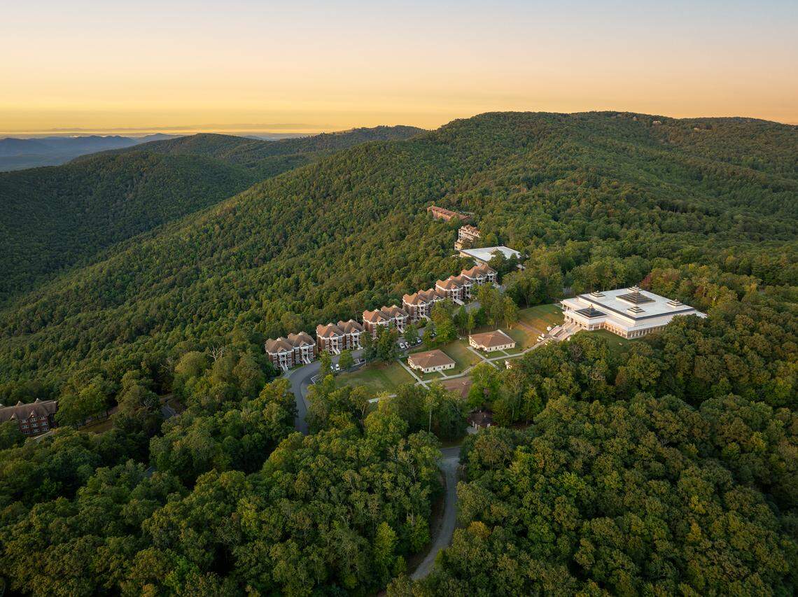 An aerial photograph taken during golden hour showcases an architectural complex situated on a ridge line surrounded by densely forested mountains. The buildings include a large, modern white structure, several clustered townhome-style residences, and smaller cabins, all nestled among the trees under a pale yellow and blue evening sky.