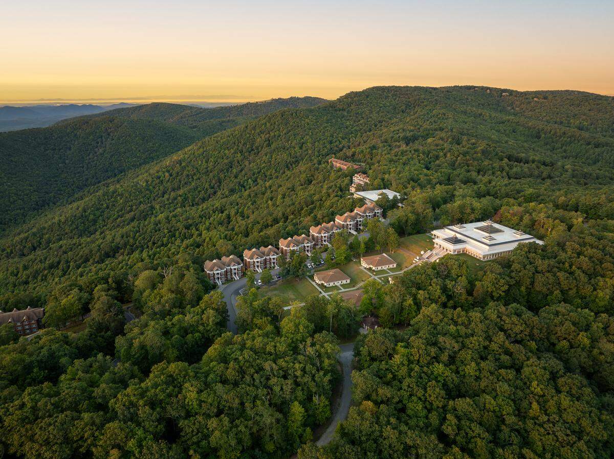 An aerial photograph taken during golden hour showcases an architectural complex situated on a ridge line surrounded by densely forested mountains. The buildings include a large, modern white structure, several clustered townhome-style residences, and smaller cabins, all nestled among the trees under a pale yellow and blue evening sky.