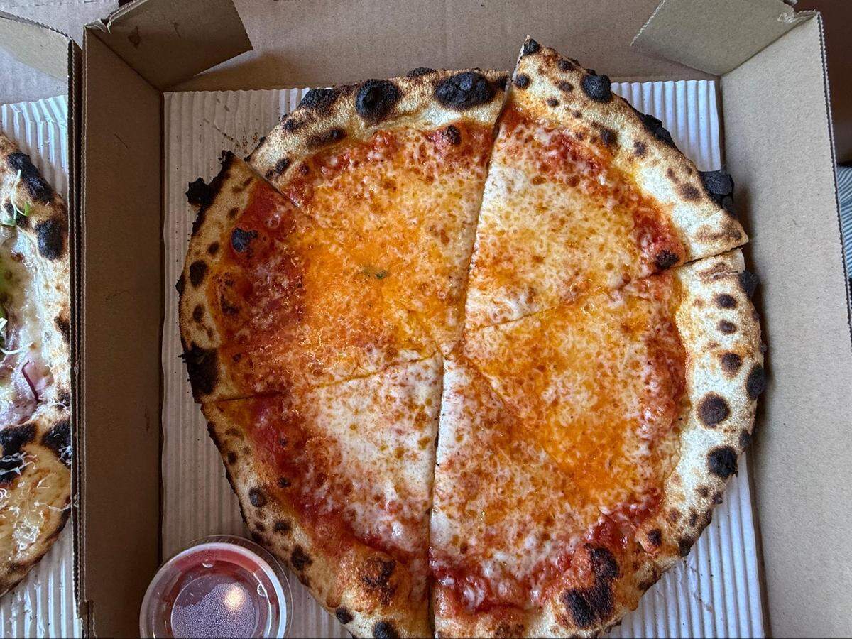 An overhead, close-up shot of an artisan-style cheese pizza in an open brown cardboard box. The pizza has a thick, bubbly, dark-charred sourdough crust and is cut into six slices. A small plastic container of red dipping sauce is visible in the bottom left corner, and a portion of another pizza is seen in an adjacent box.