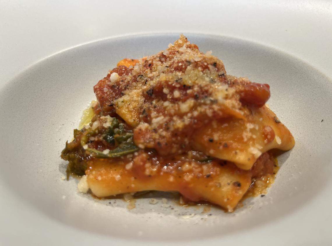 A close-up, high-angle shot of a pasta dish served in a round, light-colored bowl. The dish consists of several large, folded pasta pieces, possibly paccheri or manicotti, filled with a green mixture. They are covered in a rich, chunky red tomato sauce and generously topped with a sprinkle of grated white cheese and black pepper. The pasta is a vibrant, appealing orange-red color from the sauce. The bowl is set against a blurred light background.
