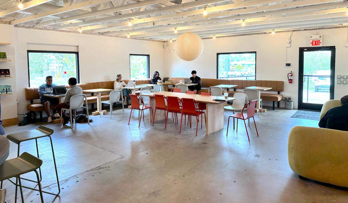 The spacious interior of a coffee shop with a minimalist aesthetic. Natural light fills the room, highlighting the white walls, exposed ceiling beams, and polished concrete floor. Patrons are scattered throughout, sitting on brown banquettes and at a central communal table with bright red-orange chairs, most focused on their laptops. A large, spherical paper lantern hangs from the ceiling.