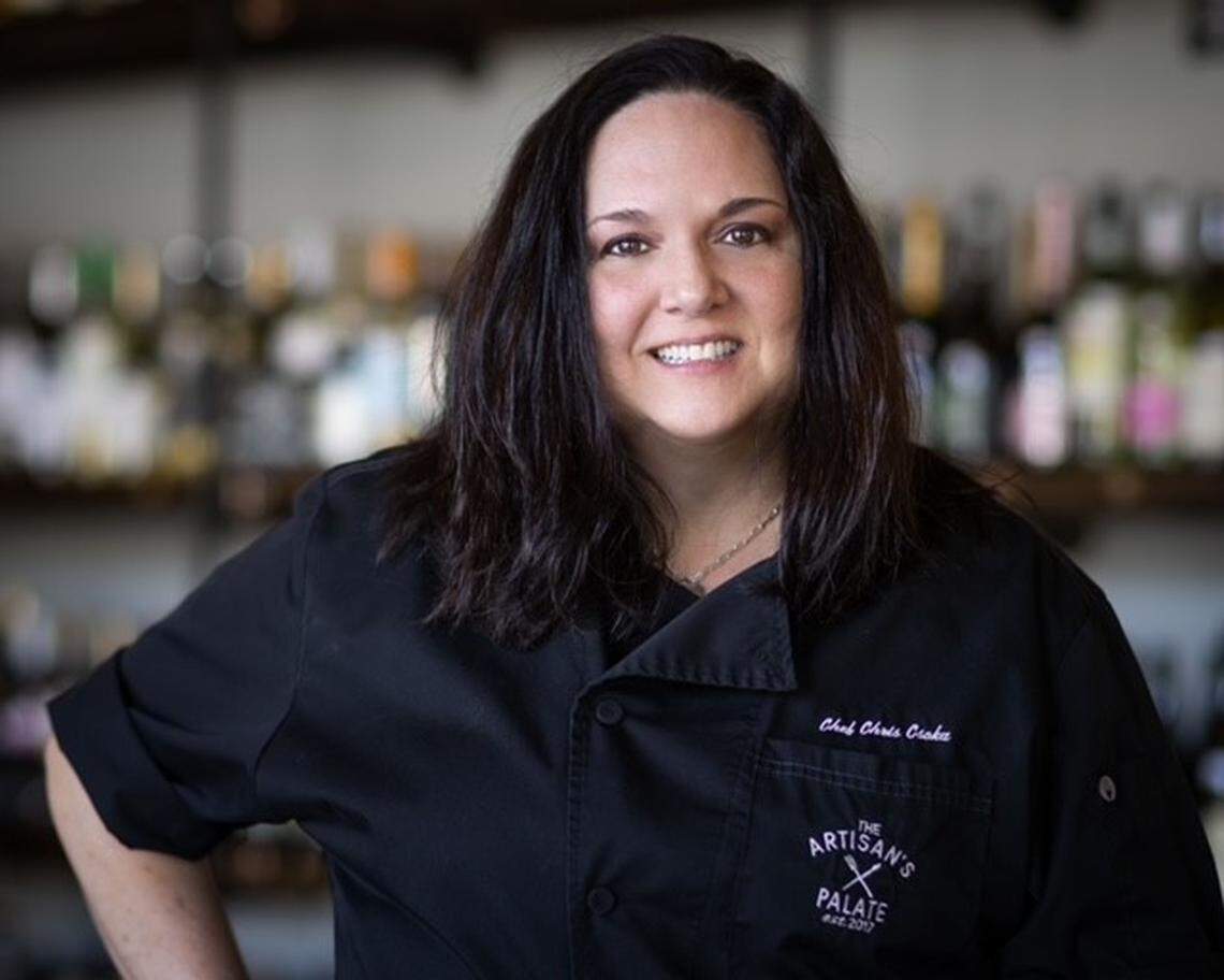 Chef Chris Clarke poses for a professional headshot. She has dark hair, is smiling and wears a black chef’s coat embroidered with her name and the logo for The Artisan’s Palate. The background is artfully blurred, showing what appears to be a well-stocked bar or wine collection.