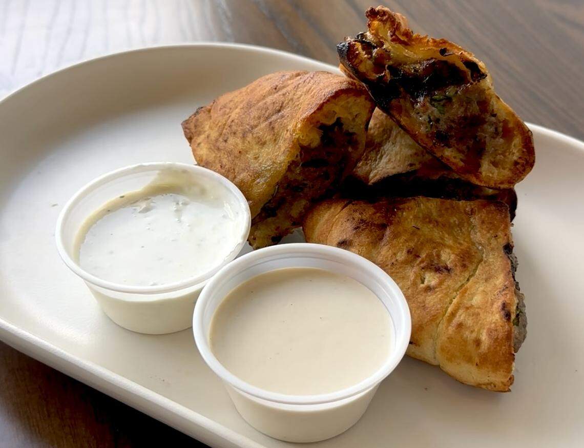 A top-down shot of a dish with fried pastries and two dipping sauces on a white, oval plate. The pastries are golden-brown and flaky, with a savory filling visible in the one that is cut open. Two small white plastic cups of sauce sit next to the pastries: one is a white, creamy sauce with herbs, and the other is a lighter, beige-colored sauce. The plate rests on a wooden table.