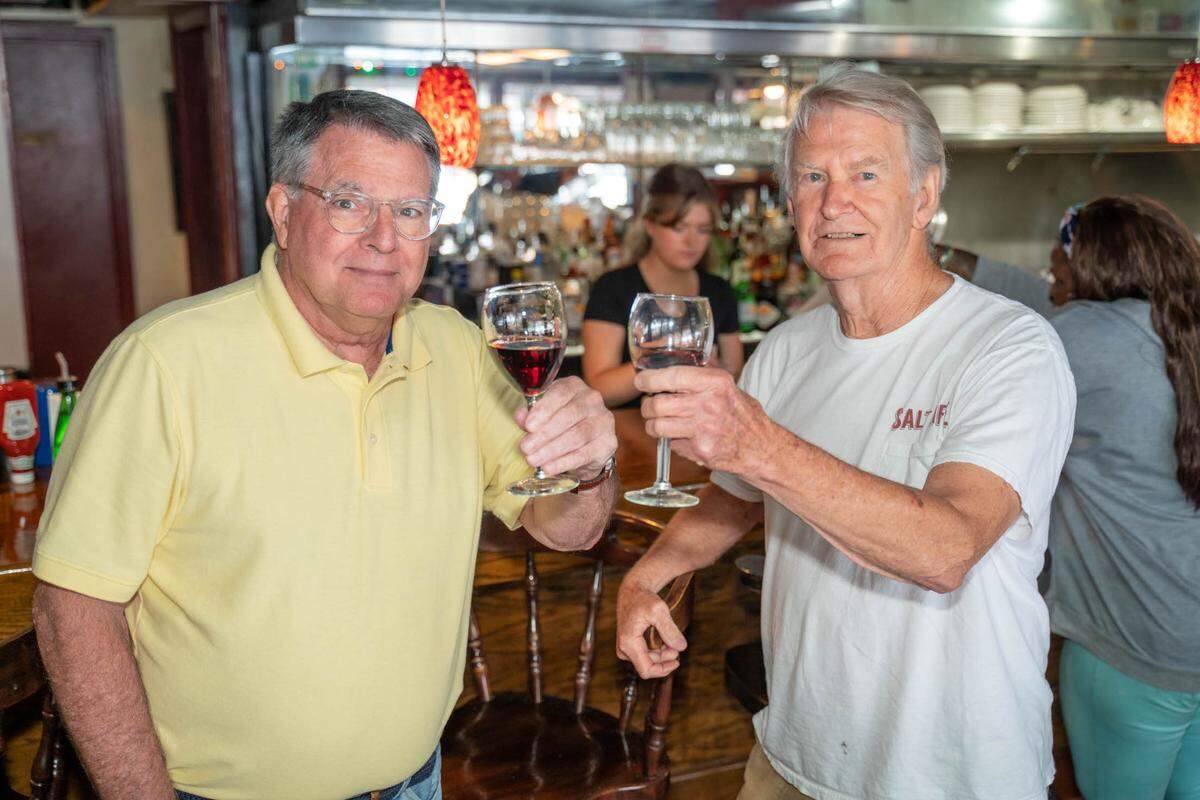 Inside a busy restaurant, two smiling people pose for the camera at the bar. They are both holding up a glass of red wine in a celebratory toast. In the background, a bartender is working, and glasses hang from a rack.