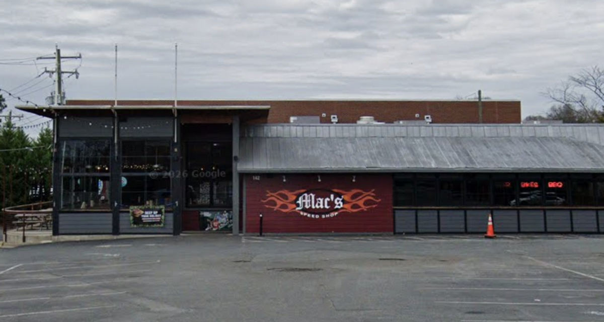 The exterior of Mac’s Speed Shop is pictured, showcasing a low-profile building with a large asphalt parking lot in the foreground. The storefront features a red wall section adorned with the restaurant’s name in stylized white text bracketed by orange flames. Large glass windows, a metal roof, and an outdoor patio seating area to the left complete the establishment’s casual, industrial look under an overcast sky.