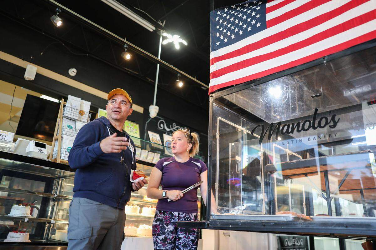 A low-angle shot captures a bakery owner in a yellow cap talking with a worker holding metal tongs inside Manolo’s Latin Bakery. A large American flag hangs prominently in the upper right, over a glass display case that reflects the bakery’s name.