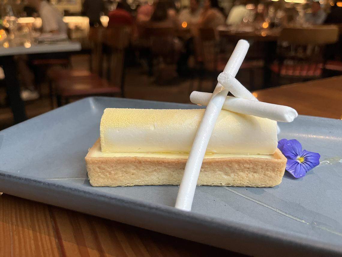 A plated dessert on a gray, rectangular plate. A rectangular tart with a golden-brown crust and a creamy filling is topped with a smooth, cylindrical yellow piece of mousse or cake. Two white, twisted meringue sticks are propped against the dessert, which is also garnished with a single purple edible flower. The plate rests on a wooden table, and the background is a blurred view of a restaurant.