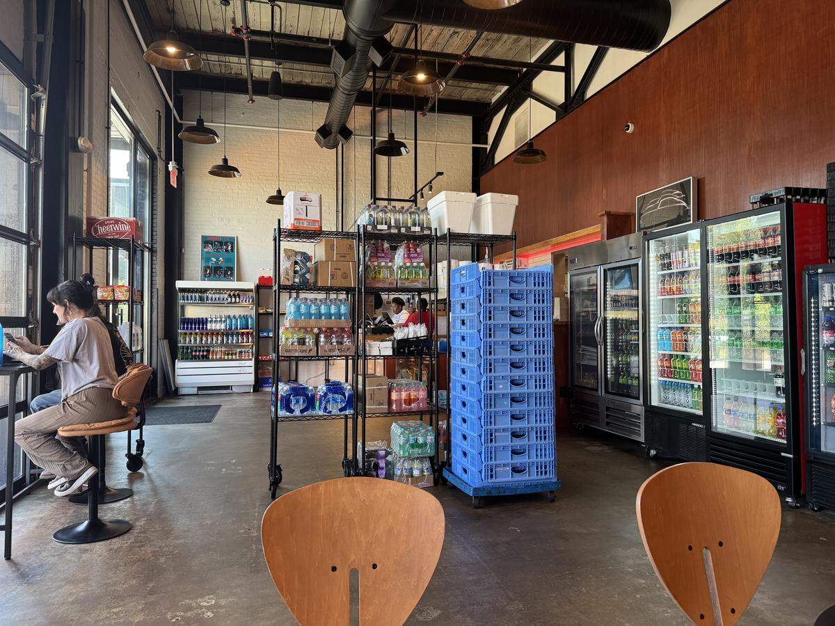 The interior of a market with exposed brick and ductwork, a large window, a variety of bottled drinks in refrigerators, and a couple of people sitting at a counter on the left. The foreground shows the backs of two empty wooden chairs.