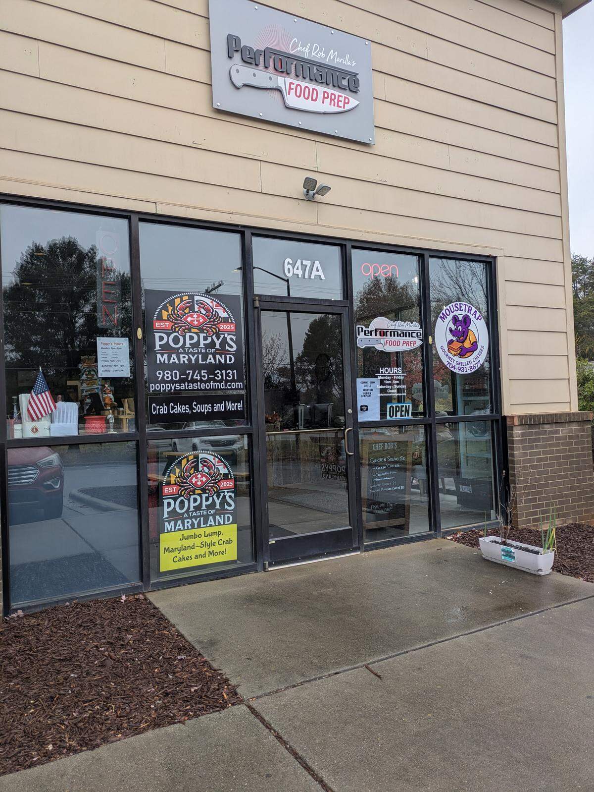 The storefront of a building containing multiple businesses: Poppy’s Maryland (Crab Cakes, Soups and More), Performance Food Prep (Chef Rob Marietta’s), and Mousetrap Grille. The entrance door is labeled “647A” and the window displays an “OPEN” sign.