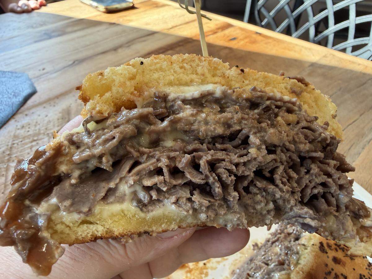 A close-up cross-section view of a gourmet roast beef sandwich held in a person’s hand. The interior reveals a thick stack of many thin layers of tender, juicy beef mixed with melted white cheese and a rich brown gravy. The bun has a golden, toasted interior and a visible seeded crust.