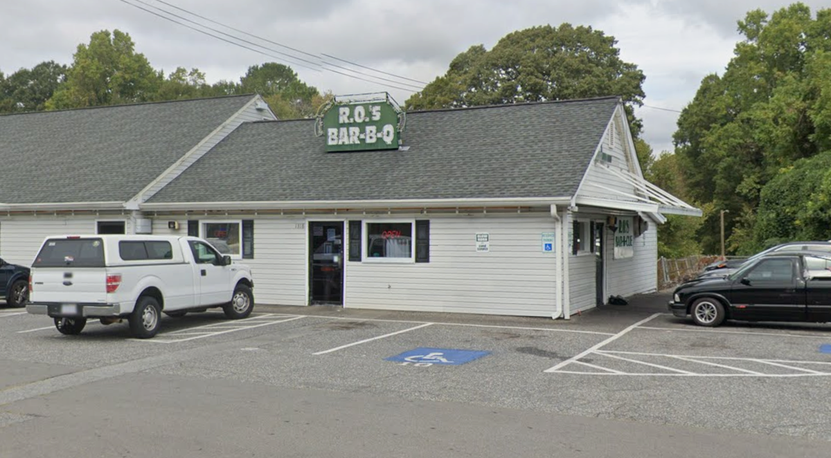 A white, single-story building with a grey shingled roof houses “R.O.’S BAR-B-Q”. The restaurant has a sign on the roof and a paved parking lot in front with several cars, including a white pickup truck. Green trees are visible in the background under an overcast sky.