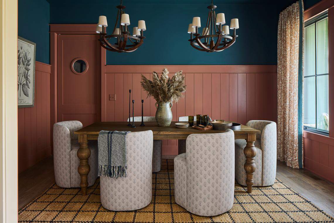 A striking dining room featuring a bold color palette of deep teal on the upper walls and ceiling and a matte terracotta or dusty rose color on the wainscoting and trim. The wainscoting is composed of vertical shiplap paneling. A long, rectangular wooden table is centered on a natural woven rug with a diamond pattern. The table is surrounded by six upholstered, swiveling barrel chairs in a pale, patterned fabric. Two rustic, multi-light wooden bead chandeliers hang above the table. A porthole window is visible on the wall, and a piece of framed botanical artwork is on the left wall.