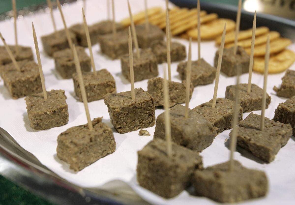 Close-up of bite-sized, square samples of liver mush or liver pudding, each topped with a wooden toothpick, arranged on a white paper-lined platter. Plain round crackers are visible in the background.