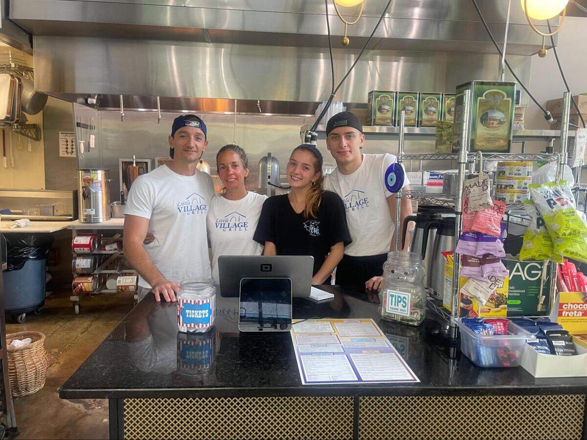 A portrait of four family members at Little Village Grill. They are standing behind a dark granite counter that holds a point-of-sale system and tip jar. The professional kitchen with stainless steel equipment is visible in the background.