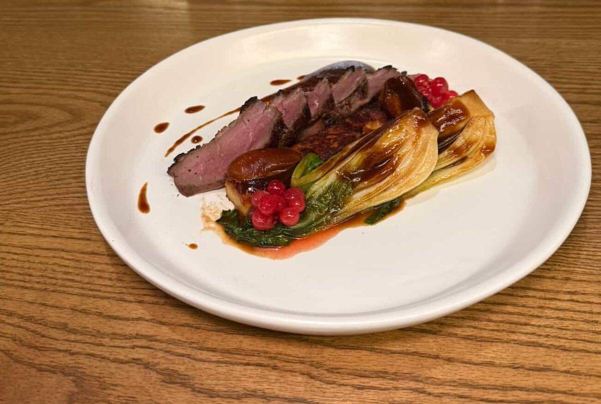 A close-up of the Dry Aged Magret Duck entree at Restaurant Constance. Slices of duck breast, cooked medium-rare with a dark, crispy skin, are fanned out next to a glistening, seared half of bok choy. The dish is garnished with a cluster of vibrant red currants and a drizzle of dark sauce on a modern white plate.