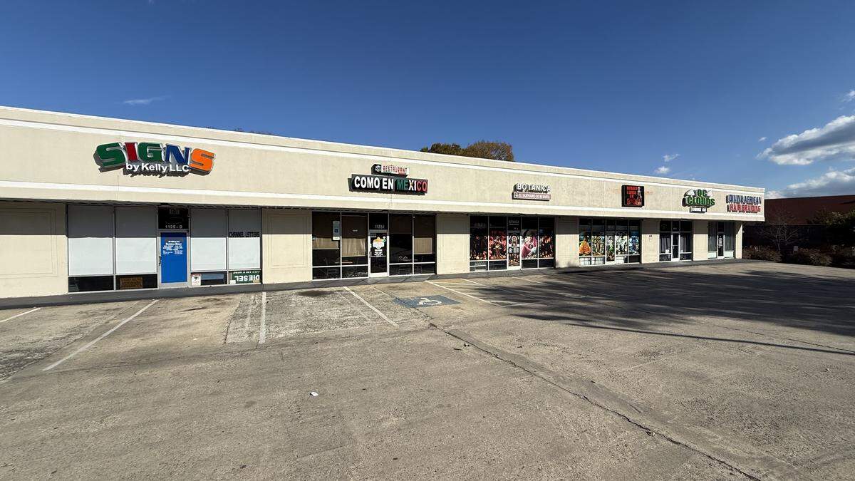 A daytime, outdoor wide-angle shot of a strip mall storefront. Visible businesses from left to right include “SIGNS by Kelly LLC,” a business in a white-front unit with the name obscured, and “COMO EN MEXICO,” “BOTANICA LA MADRINA,” and several other storefronts with brightly colored signage. The businesses are housed in a long, beige building with a flat roof. A large, empty concrete parking lot is in the foreground with accessible parking spaces marked.