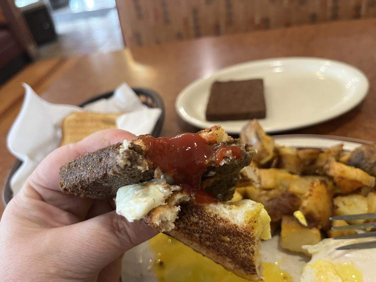 A close-up of a hand holding a half-eaten liver mush and egg sandwich, with a dollop of bright red ketchup on top. The plate below shows home fries and a runny egg yolk.