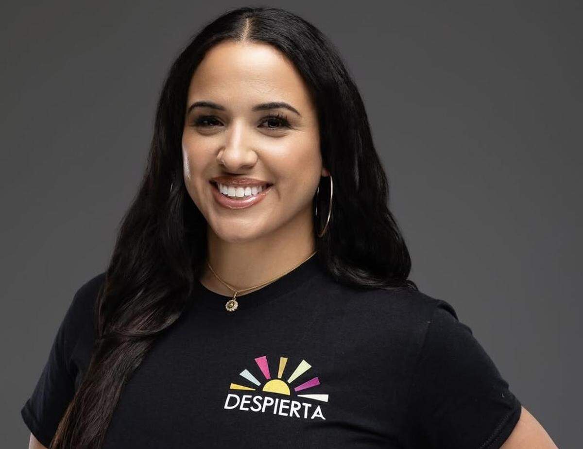 A close-up studio portrait of a smiling person with long dark hair and hoop earrings, wearing a black t-shirt that features a colorful rising sun logo above the text “DESPIERTA.”