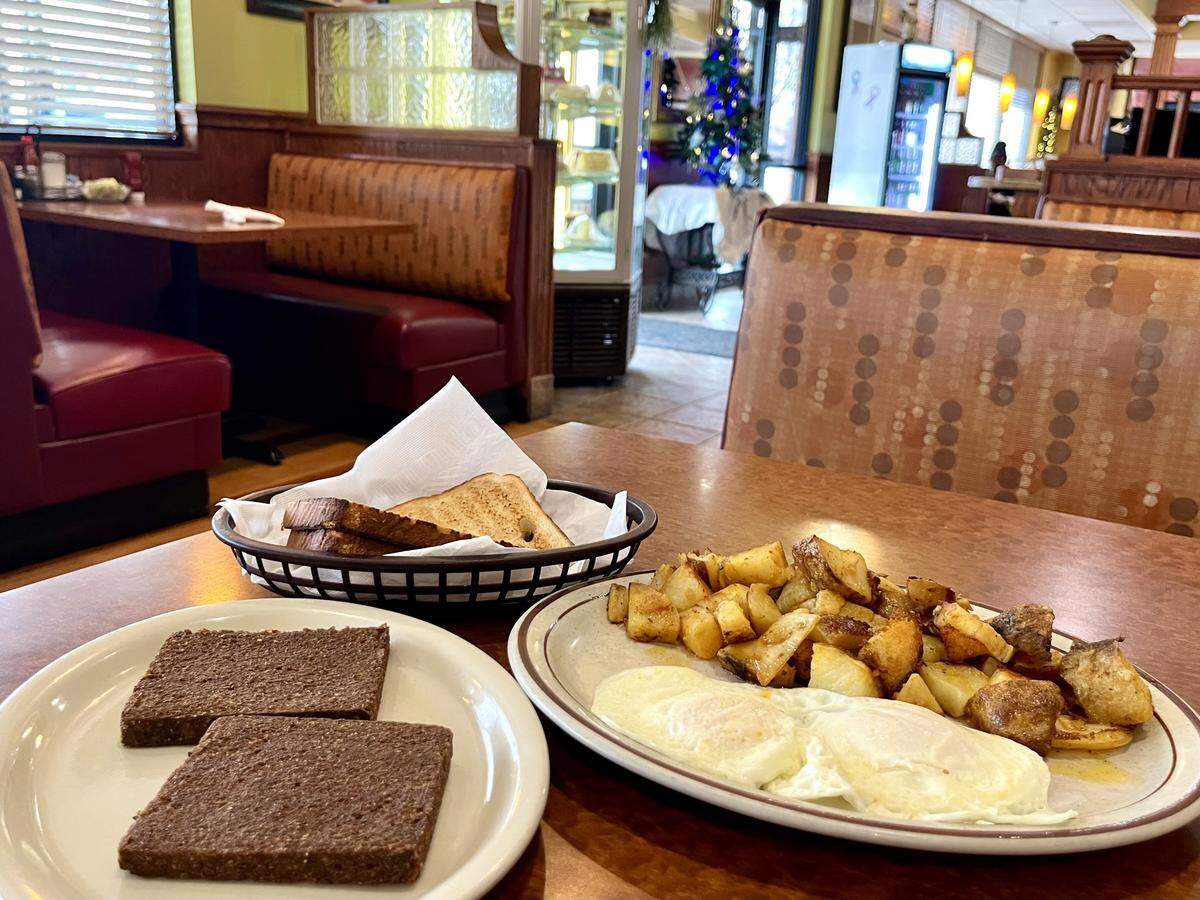 A wide shot of a breakfast setup in a cozy diner. On the right, a plate holds two sunny-side-up eggs and a pile of home fries. On the left, a smaller plate holds two square slices of cooked livermush. A basket of toast is between the plates. The background features warm-toned vinyl booths and a decorated Christmas tree.