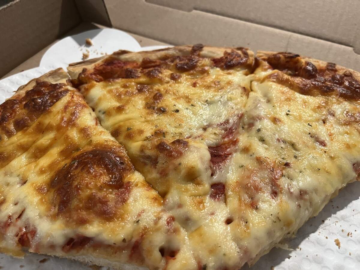 A close-up shot of three slices of a cheese pizza, still in a cardboard takeout box. The pizza has a golden-brown, bubbly layer of melted cheese, with hints of red tomato sauce visible underneath. The crust is hearty, and the cheese is sprinkled with what appears to be dried oregano or another herb. The pieces of pizza are slightly greasy and look freshly made.