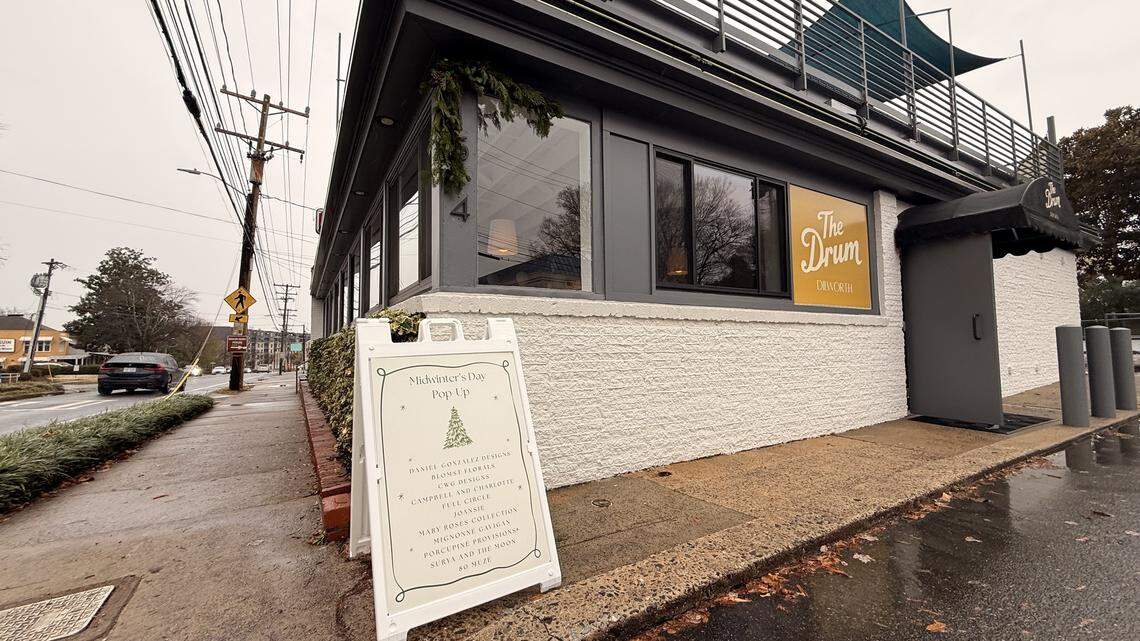 The exterior of “The Drum Dilworth” building on a cloudy day, featuring a white textured facade and large dark-framed windows. An A-frame sandwich board sign on the sidewalk advertises a “Midwinter’s Eve Pop-Up” event with a list of vendors. The building has a dark grey trim, and a small area with an outdoor railing is visible on the roof.