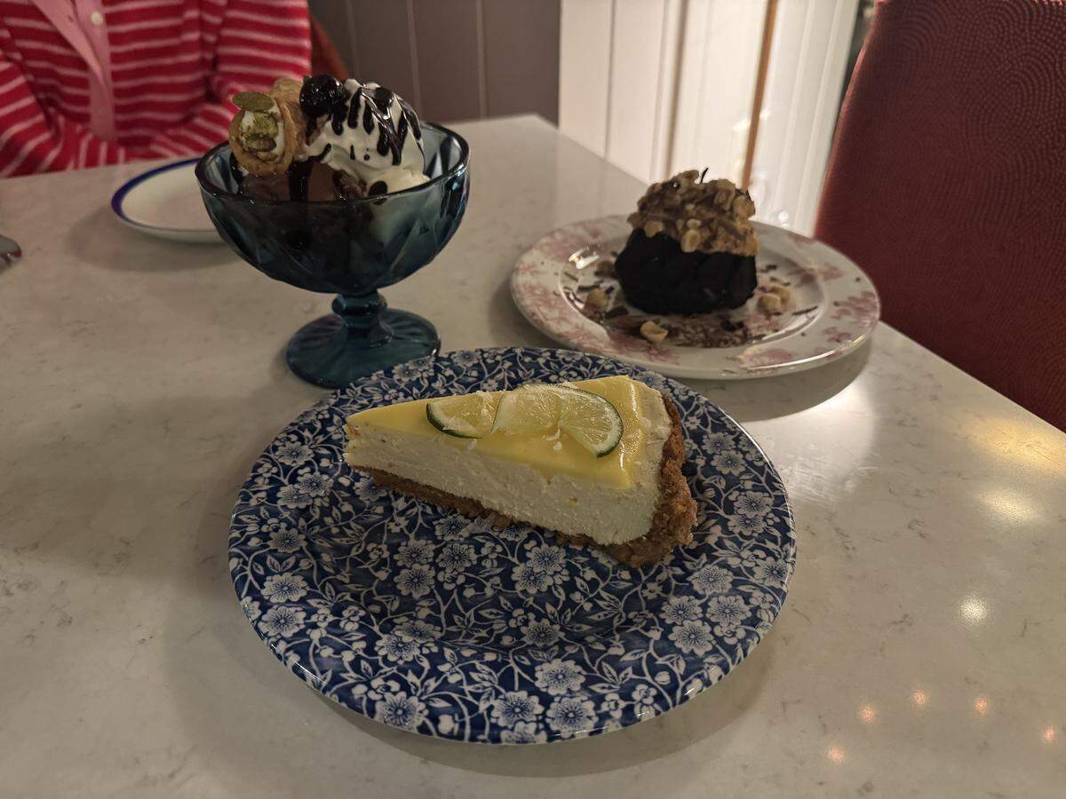 Three desserts arranged on a white marble table. In the foreground, a slice of lemon or lime tart sits on a blue-and-white floral plate, topped with translucent citrus wheels. To the left, an ice cream sundae with chocolate sauce, whipped cream, a cherry, and a mini cannoli is served in a blue glass goblet. In the background, a dark chocolate lava cake or dome dessert sits on a pink-and-white floral plate.