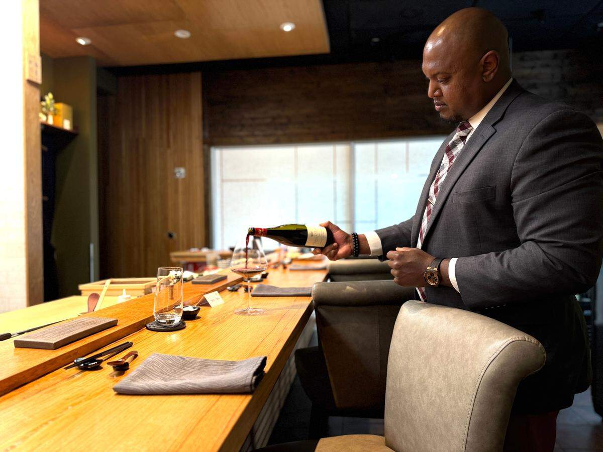 A well-dressed bald man in a dark suit with a patterned tie is pouring red wine from a green bottle into a wine glass on a light wooden bar counter. He is focused on the task, with a watch visible on his left wrist. The bar counter is set with several place settings, including folded napkins, glasses, and small dishes. In the blurred background, there’s a window and a modern restaurant interior with wooden accents and various shelves.