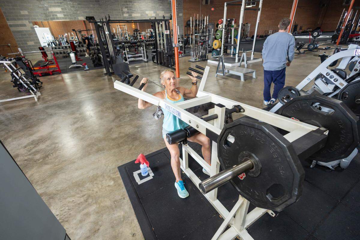 A person with a bright blue top and black shorts sits on a white, plate-loaded shoulder press machine inside a gym. They are actively performing a repetition, looking up toward the camera, and the gym floor and other exercise equipment are visible in the background.