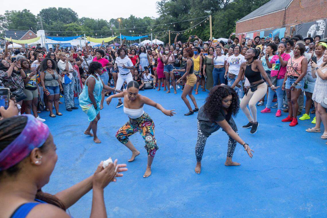 A vibrant outdoor community event where several women are dancing in the center of a large blue-painted courtyard. A large, diverse crowd of people stands in a circle around them, many of whom are smiling, clapping, and recording the performance on their phones. The scene is set outdoors during the day with string lights hanging overhead and brick buildings in the background.