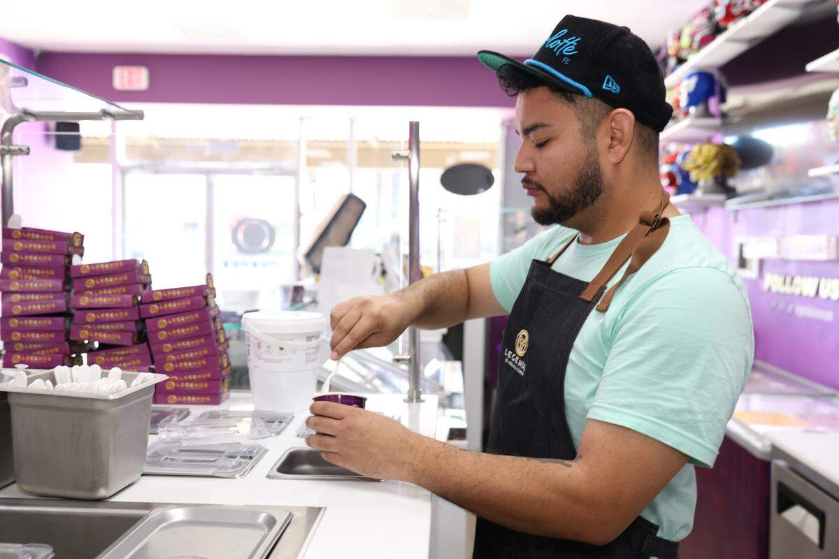 An eye-level, medium shot shows an ice cream shop worker from the side, standing behind a white service counter while wearing a mint-colored t-shirt, a black apron, and a backward black baseball cap. The worker is focused on stirring something in a small purple cup with a white spoon. The background shows stacks of purple boxes and the restaurant’s bright interior and entrance.