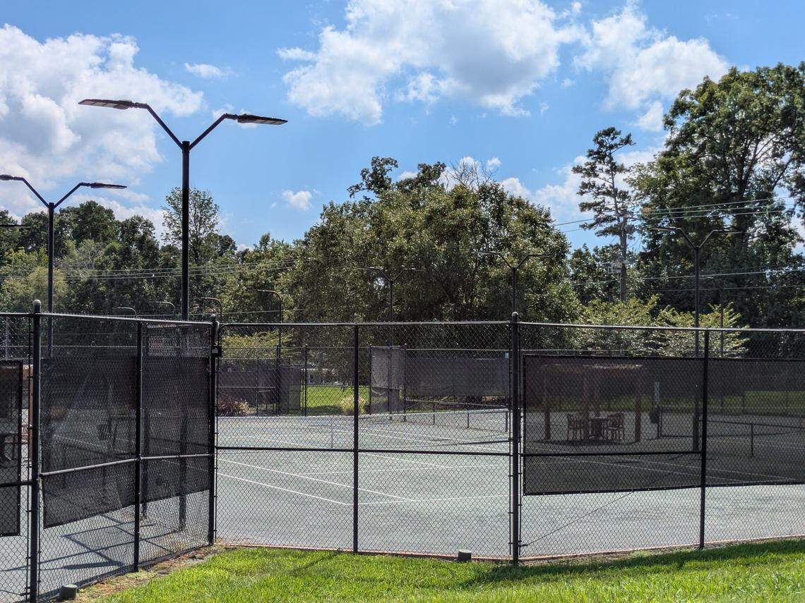 A tranquil outdoor tennis court, seen from outside its black chain-link fence. The pale green court is empty, with modern light fixtures standing by for evening play. In the background, a lush treeline meets a bright blue sky dotted with clouds.
