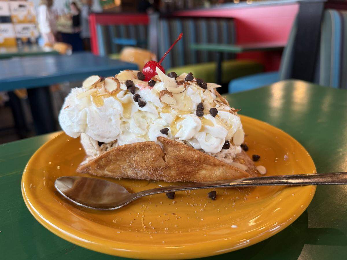 A dessert taco featuring a crispy, cinnamon-dusted shell filled with ice cream. It is piled high with whipped cream, drizzled with caramel sauce, and topped with sliced almonds, chocolate chips, and a maraschino cherry.