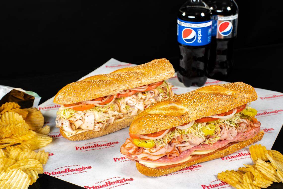 A close-up, studio-style shot of two large, appetizing PrimoHoagies Italian specialty sandwiches on sesame seed rolls, resting on branded butcher paper. They are filled with deli meats, cheese, lettuce, tomatoes, and onions. To the left are ruffled potato chips, and in the background are two bottles of Pepsi.