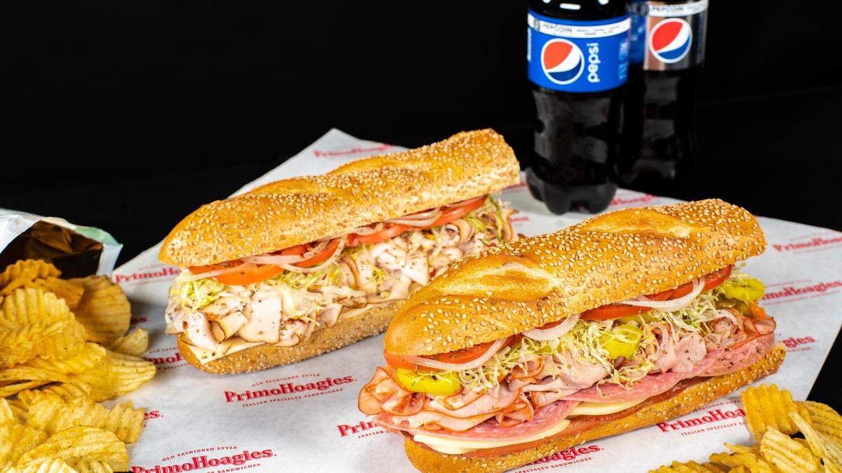 A close-up, studio-style shot of two large, appetizing PrimoHoagies Italian specialty sandwiches on sesame seed rolls, resting on branded butcher paper. They are filled with deli meats, cheese, lettuce, tomatoes, and onions. To the left are ruffled potato chips, and in the background are two bottles of Pepsi.