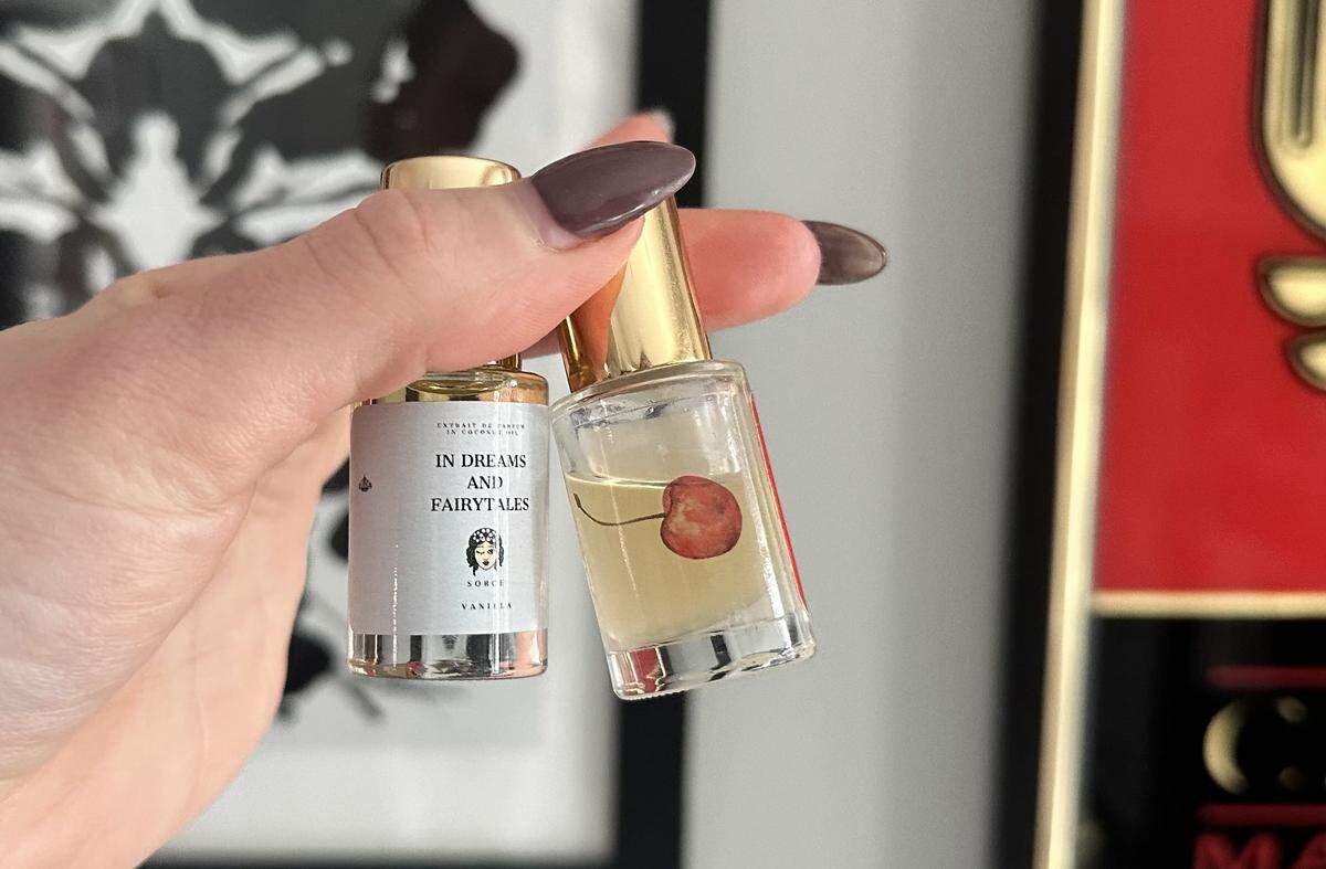 A close-up shot of a hand with mauve-colored fingernails holding two small perfume oil bottles. The bottle on the left has a white label that reads “IN DREAMS AND FAIRYTALES” with a small illustration of a woman’s face. The bottle on the right is clear, containing a light yellow liquid and featuring a realistic illustration of a red cherry on the glass.