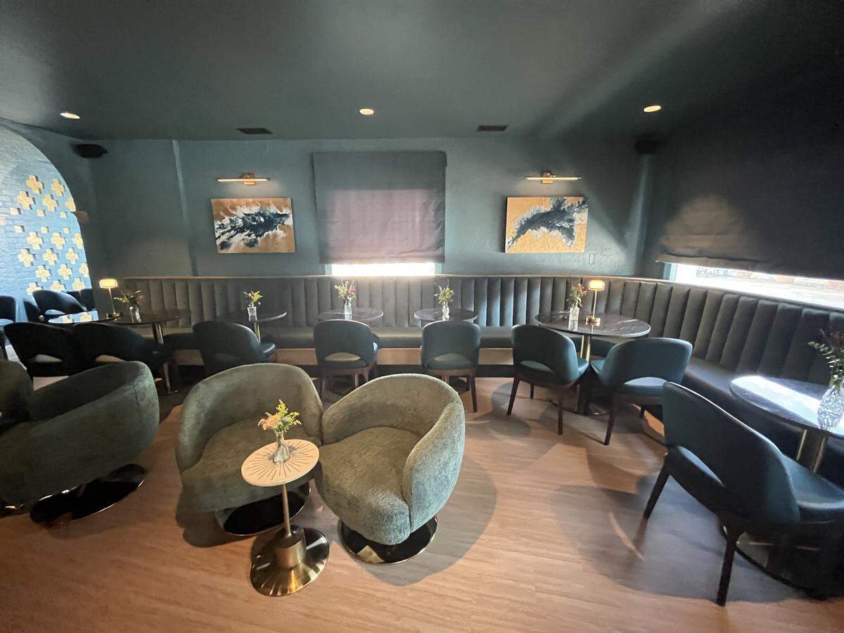 A sweeping view of an elegantly decorated lounge. Along the walls is a dark blue-green, channel-tufted banquette, while the center of the room features pairs of light green club chairs. The dark walls are hung with abstract art, and on the left, an arched nook is decorated with a pattern of gold tiles.
