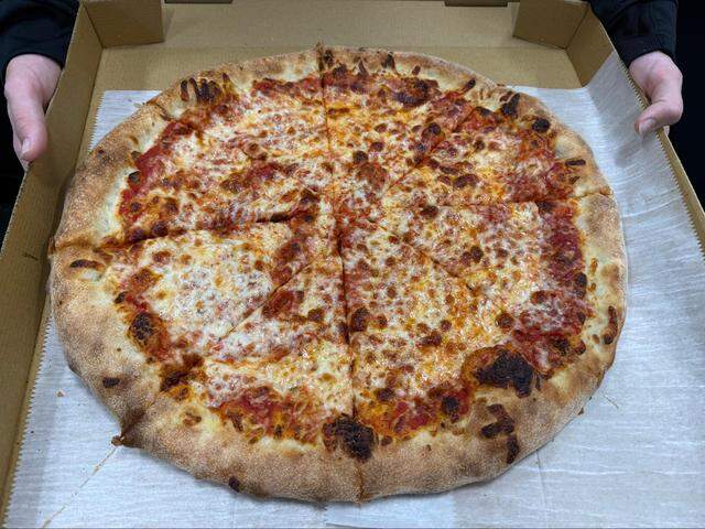 An overhead, close-up shot shows a large, circular cheese pizza in a cardboard delivery box. The pizza is cut into eight slices and rests on white parchment paper. It features a thick, golden-brown crust with several charred, dark spots around the edges. The center is covered in melted mozzarella cheese with toasted brown patches, with glimpses of red tomato sauce visible beneath the cheese. A person’s hands are visible on the left and right sides, holding the edges of the box.