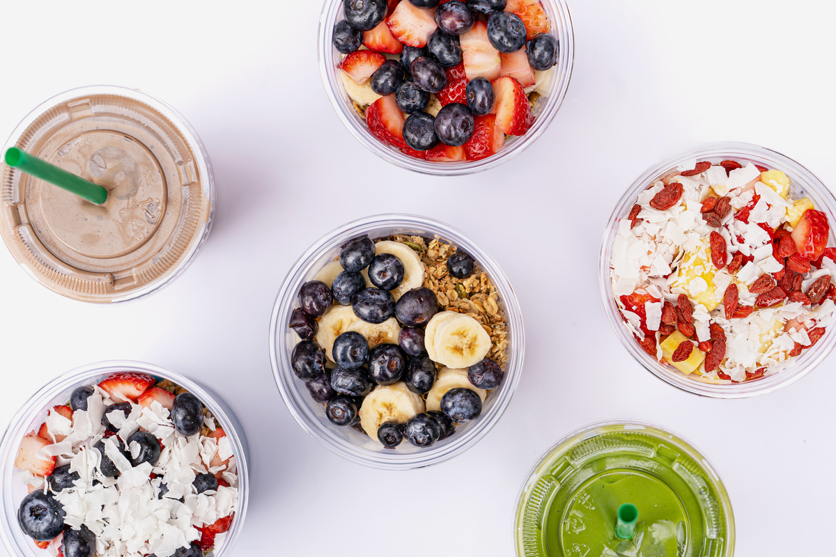 A top-down view of six South Block menu items arranged on a white surface. The spread includes four acai bowls in clear plastic cups, topped with fresh ingredients like strawberries, blueberries, sliced bananas, goji berries, and coconut flakes. Two smoothies in clear cups with green straws are also visible—one a creamy chocolate-brown color and the other a vibrant green.
