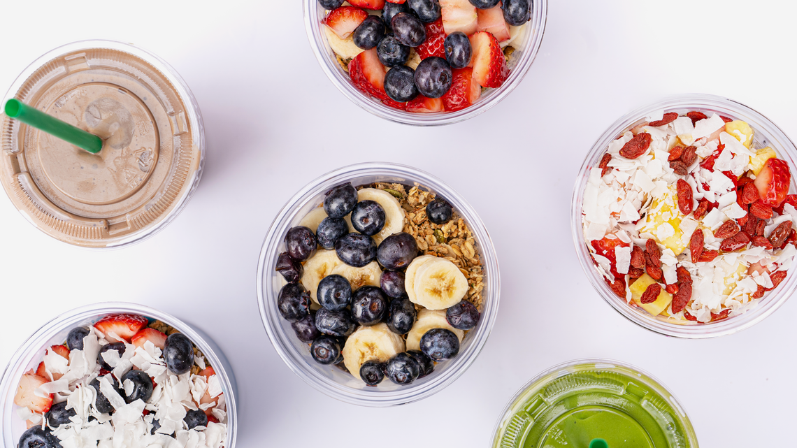 South Block is known for its smoothies, acai bowls and more. A top-down view of six South Block menu items arranged on a white surface. The spread includes four acai bowls in clear plastic cups, topped with fresh ingredients like strawberries, blueberries, sliced bananas, goji berries, and coconut flakes. Two smoothies in clear cups with green straws are also visible—one a creamy chocolate-brown color and the other a vibrant green.