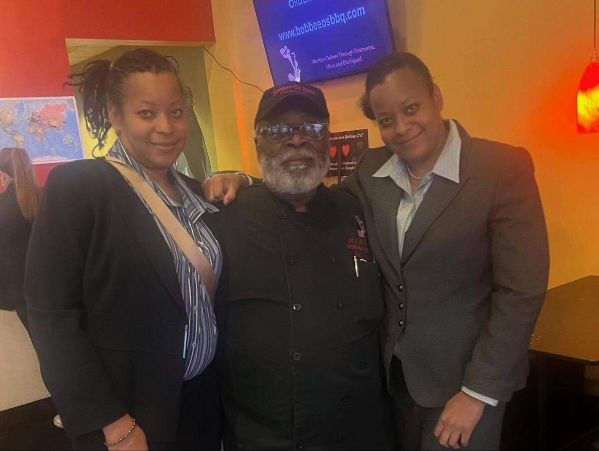 Three people standing together in a dimly lit restaurant interior. The person in the center wears a black chef’s coat and a baseball cap, flanked by two people in professional blazers. They are all looking at the camera with soft smiles.