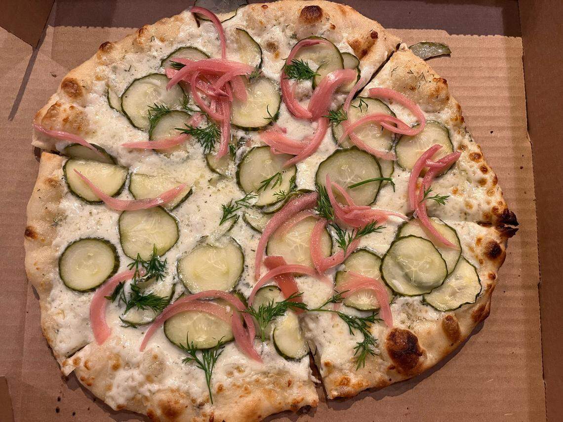 An overhead, close-up shot shows a specialty pizza in a cardboard box. The pizza has a white sauce base and is topped with numerous thin slices of pickles, pickled red onions, and fresh dill fronds, with a browned, bubbled crust.