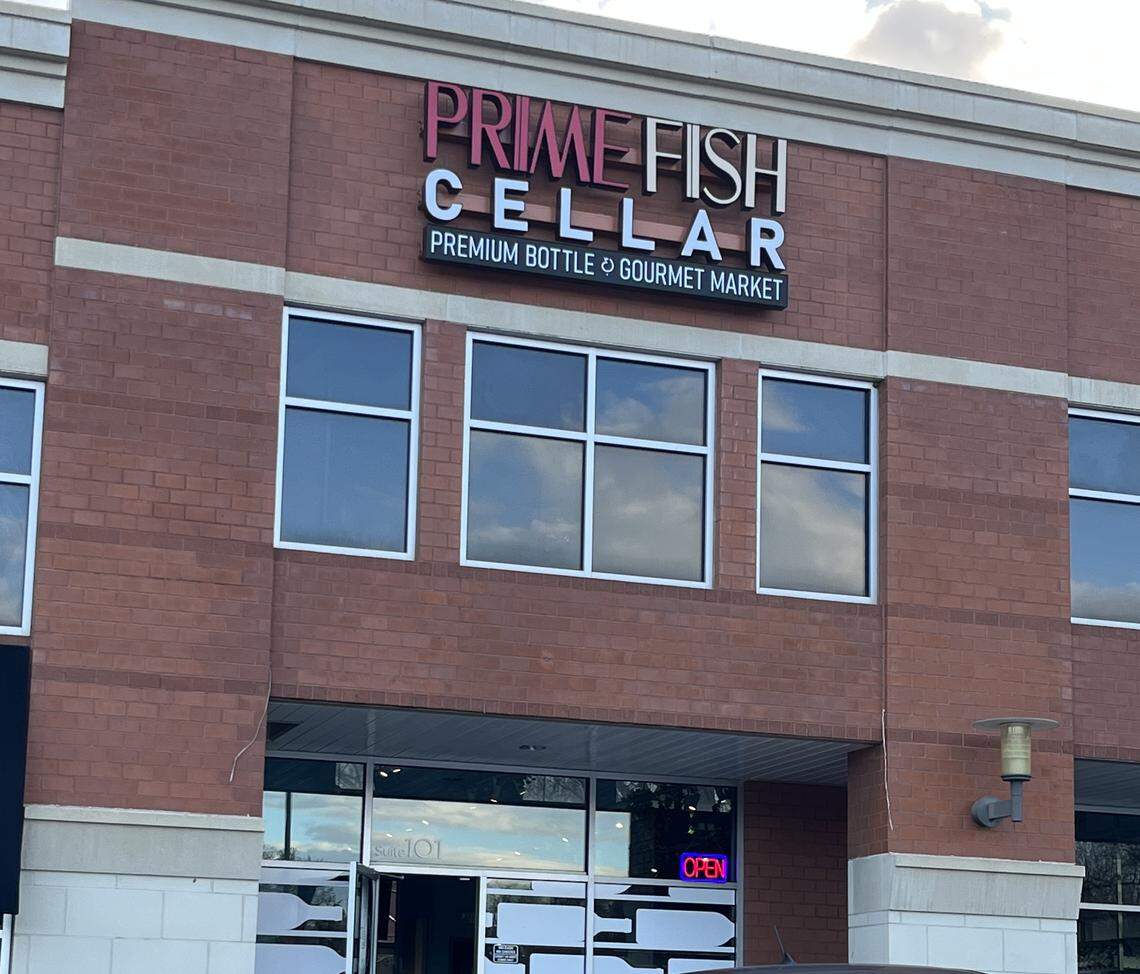 The exterior of a two-story, red brick building featuring the storefront for “Prime Fish Cellar.” The sign has “PRIME FISH” in pink letters and “CELLAR” in white. Below this, a smaller sign reads “PREMIUM BOTTLE & GOURMET MARKET.” A neon “OPEN” sign is visible in the window.