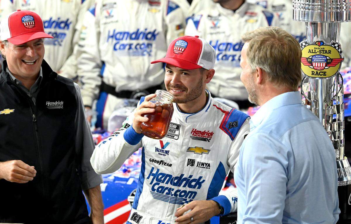 NASCAR driver Kyle Larson, center, takes a swig of moonshine in victory lane as former driver Jeff Gordon, left and Marcus Smith, CEO of Speedway Motorsports, right, look on as the team celebrate winning the NASCAR All-Star race at North Wilkesboro Speedway on Sunday, May 21, 2023.