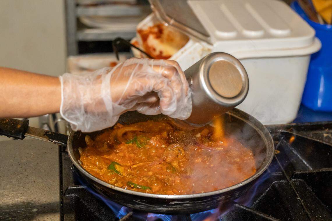 A high-detail kitchen shot focusing on the cooking process. A hand wearing a clear plastic glove pours spices from a stainless steel shaker into a pan of simmering, vibrant red stew. Flames from the gas range lick the sides of the pan, and steam rises, capturing the aromatic and intense flavors being developed in the kitchen.