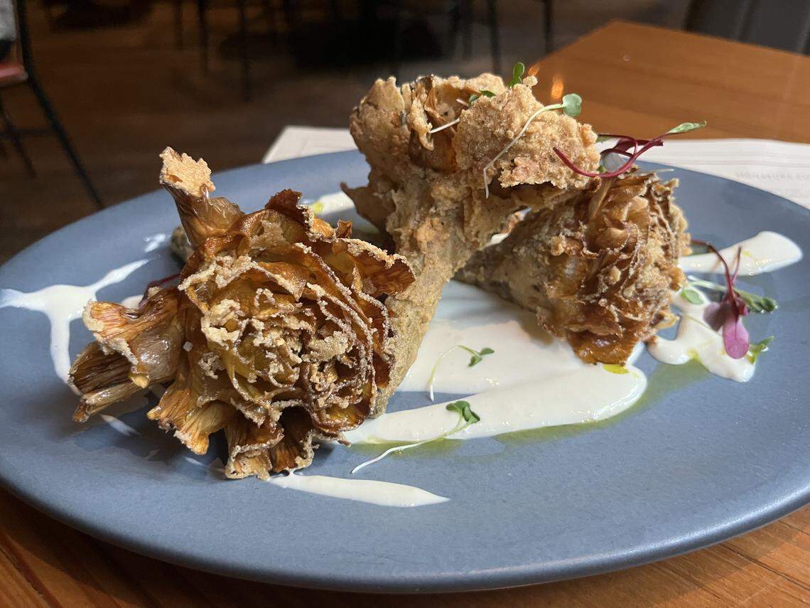A high-angle, close-up shot of a dish of fried artichoke hearts served on a gray plate. The artichoke hearts are golden-brown and crispy, with a feathery texture, and are arranged on a white, creamy sauce. The dish is garnished with small green and purple microgreens and a drizzle of olive oil.