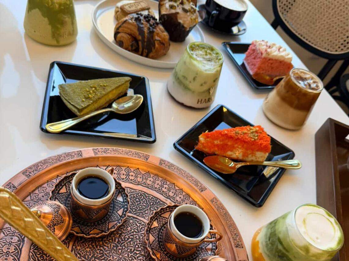 A diverse spread of desserts and beverages is arranged on a white table, including slices of cake, pastries, and layered iced drinks. Two small copper cups of dark, hot coffee rest on an ornately engraved copper tray in the foreground.
