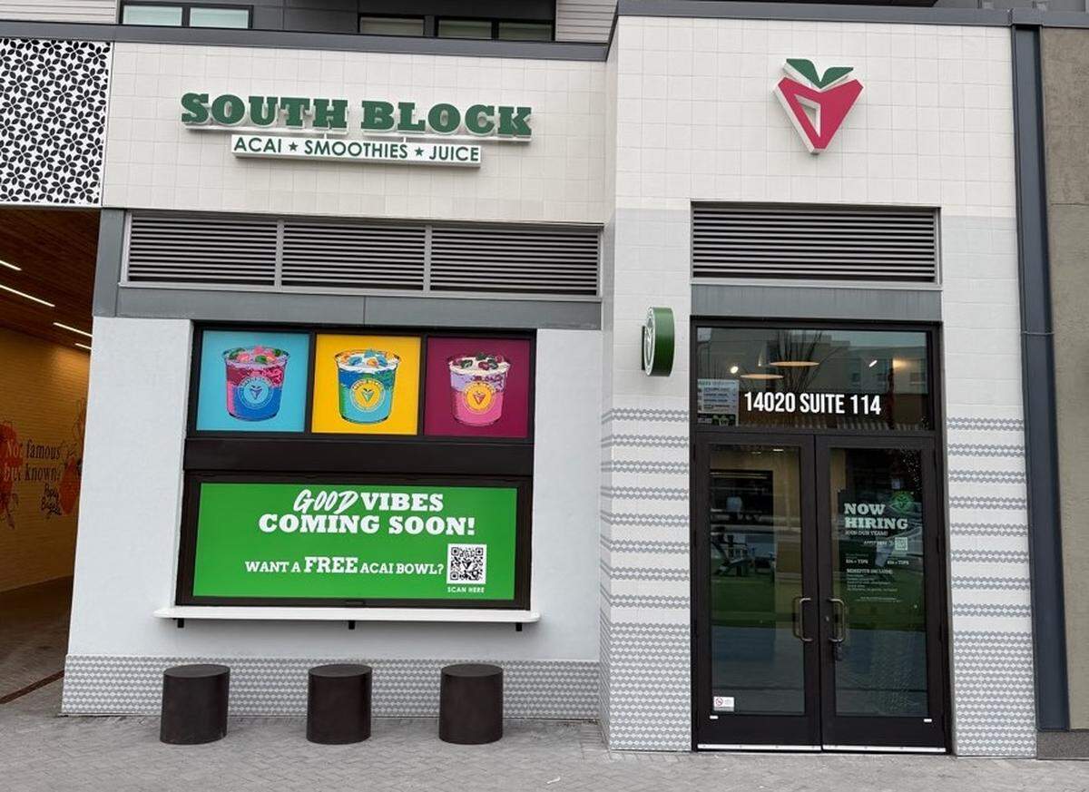 The modern exterior storefront of South Block in Ballantyne, featuring white tiled walls and a black-framed glass entrance door. Above the door, the address “14020 SUITE 114” is visible, and the company logo—a stylized pink and green fruit—is mounted on the wall. A large window display features colorful graphics of acai bowls and a green banner that reads “GOOD VIBES COMING SOON!” with a QR code. Three small cylindrical dark stools are placed on the sidewalk in front of the window.