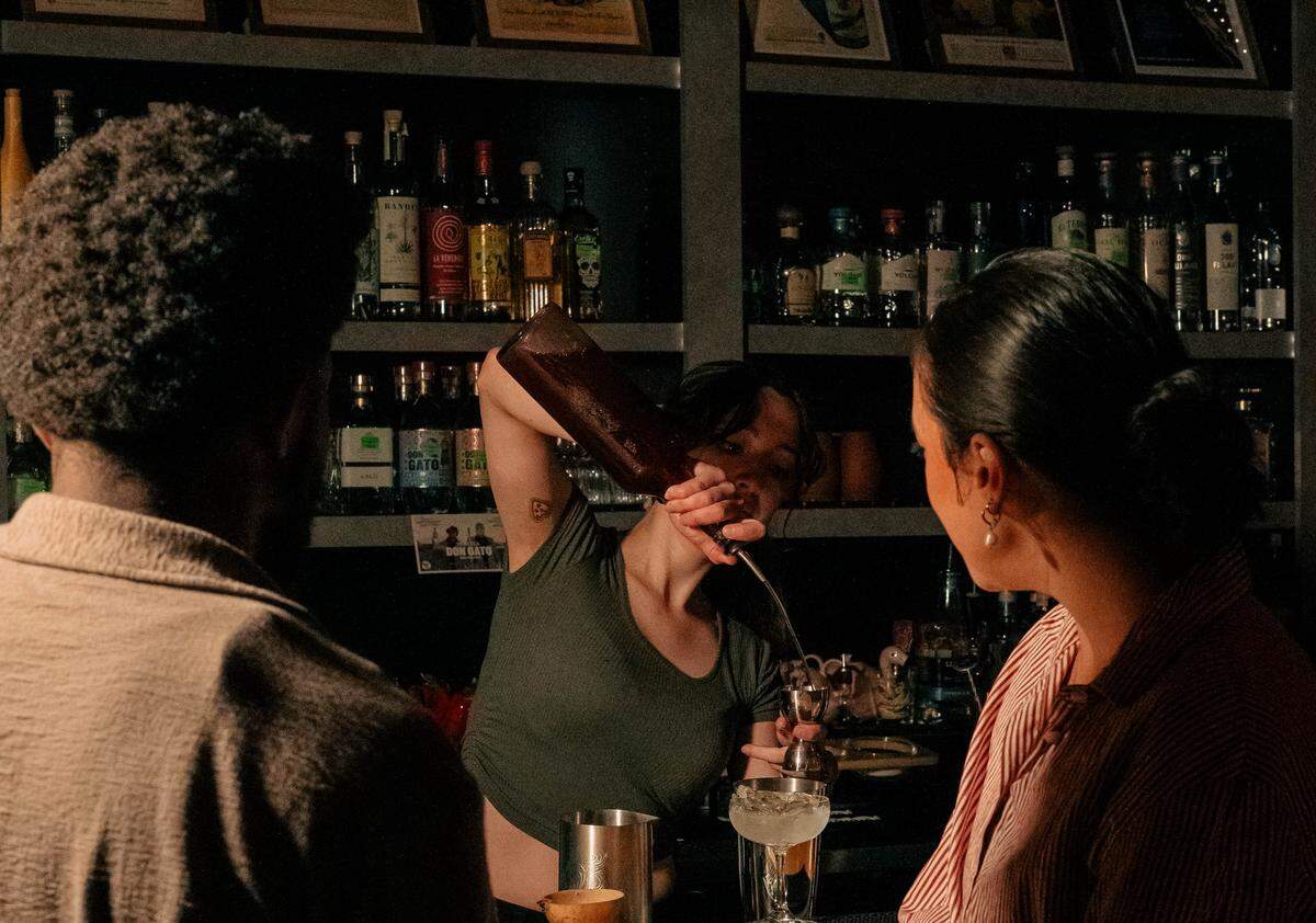 A bartender, seen between two customers whose backs are to the camera, skillfully pours a drink from a dark bottle into a mixing glass. The setting is a dimly lit, intimate bar with shelves packed with liquor bottles in the background.